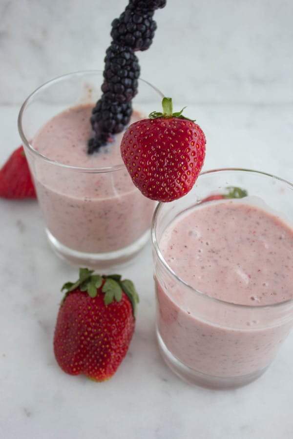 2 glasses of Banana Berry Avocado Chia Smoothie on a white kitchen counter decorated with a blackberry skewer and some fresh strawberries