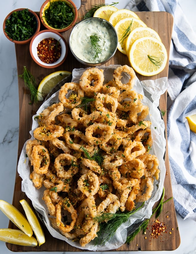 A whole plate of fried calamari served over a board with tartar sauce, lemon slices, extra chilli flakes and dill to sprinkle