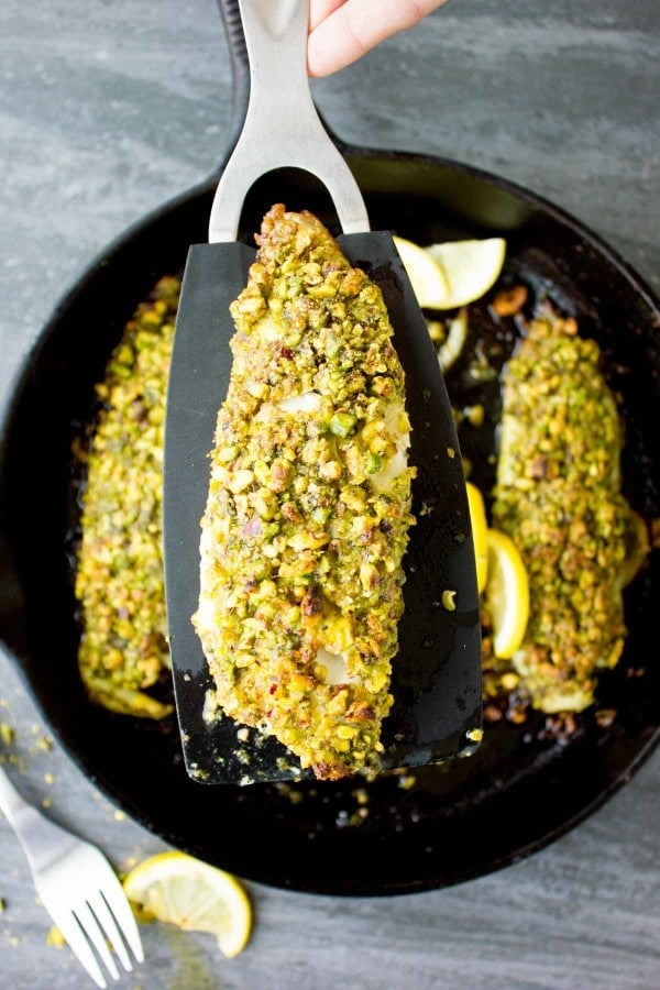 Pesto Pistácie Fish Fillet na černé kuchyňské lopatce držené nad pánví s dalšími rybími filety.