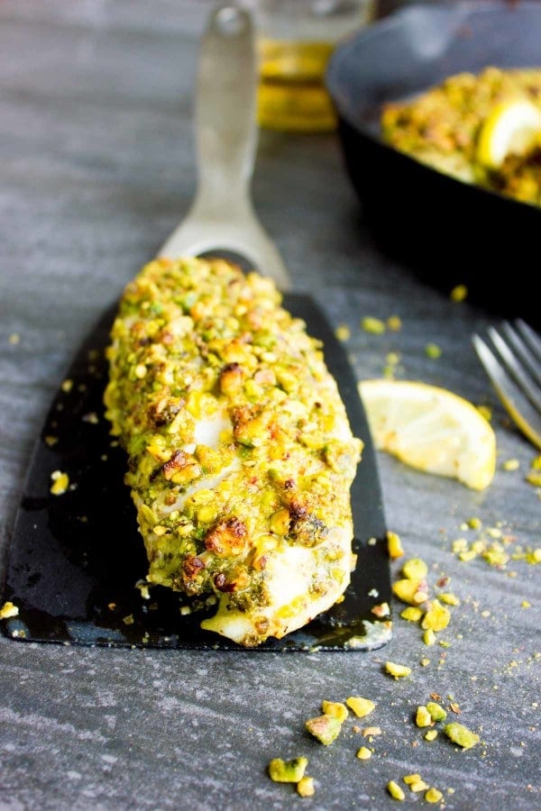 Pistachio crusted fish fillet on a black kitchen spatula with a black skillet with more fish fillets in the background.
