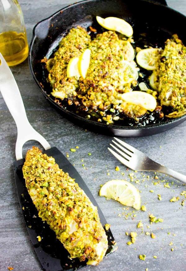 Pistachio crusted fish fillet on a black kitchen spatula placed next to a black skillet with three more fish fillets.
