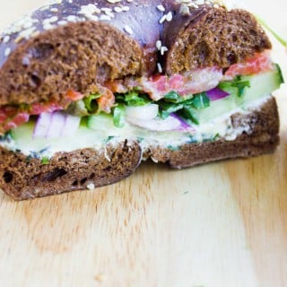 Close up of a bagel breakfast sandwich showing the layering of cream cheese, cucumbers, onions, spinach, smoked salmon and sauce in a pumpernickel bagel