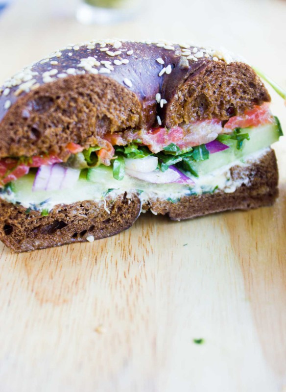 Close up of a bagel breakfast sandwich showing the layering of cream cheese, cucumbers, onions, spinach, smoked salmon and sauce in a pumpernickel bagel