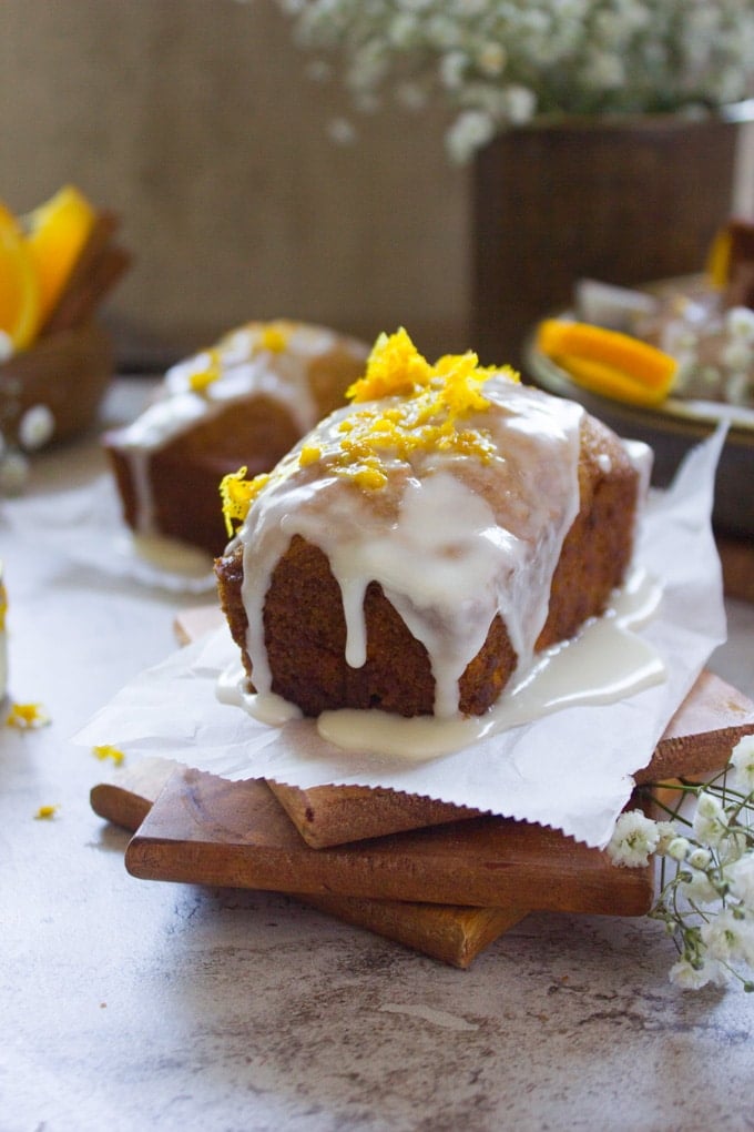 Close up of one orange carrot cake glazed and topped with orange zest over a wooden coaster.
