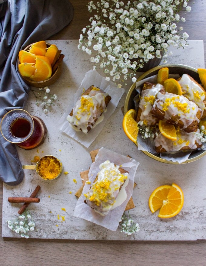 Top view showing a plate of mini carrot cakes, white flowers, a cup of tea, half oranges, cinnamon sticks