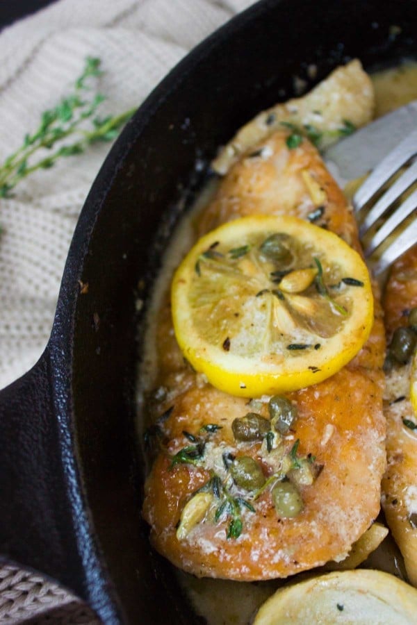 close-up of a piece of Lemon Caper Artichoke Chicken Piccata in a black skillet topped with a lemon slice