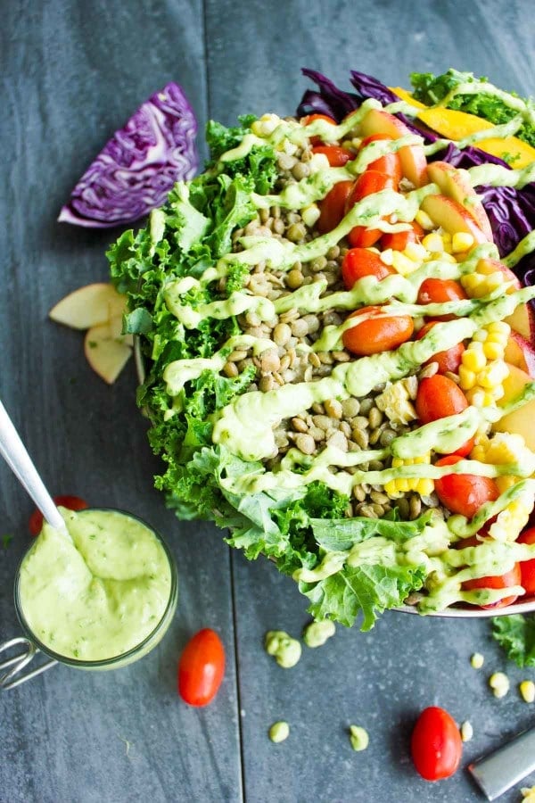 Vegan Mexican Style Lentil Detox Salad topped with fresh apples, mangoes, tomatoes and a creamy guacamole dressing served in a big salad bowl on a rustic dark wood table with a side dish of Green Goddess Dressing