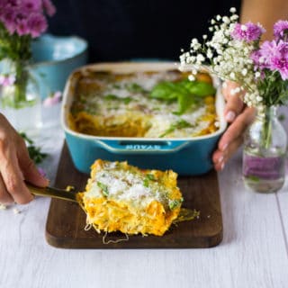 A hand holding a serving spoon serving up a slice of butternut squash lasagna on a wooden board