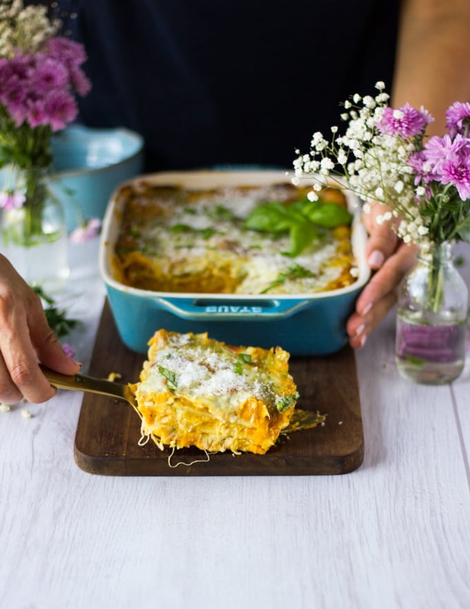 A hand holding a serving spoon serving up a slice of butternut squash lasagna on a wooden board