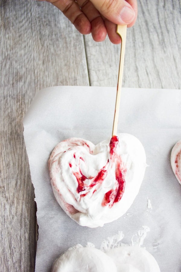 heart shaped meringue on a piece of parchment paper with a hand swirling some raspberry sauce into the meringue with a skewer