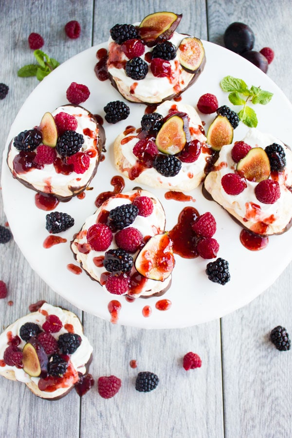 Chocolate Dipped Mini Pavlova drizzled with raspberry sauce and decorated with fresh berries and fig slices on a white cake stand