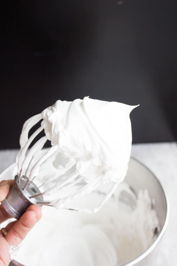 a whisk with stiff egg white meringue held over a bowl with meringue