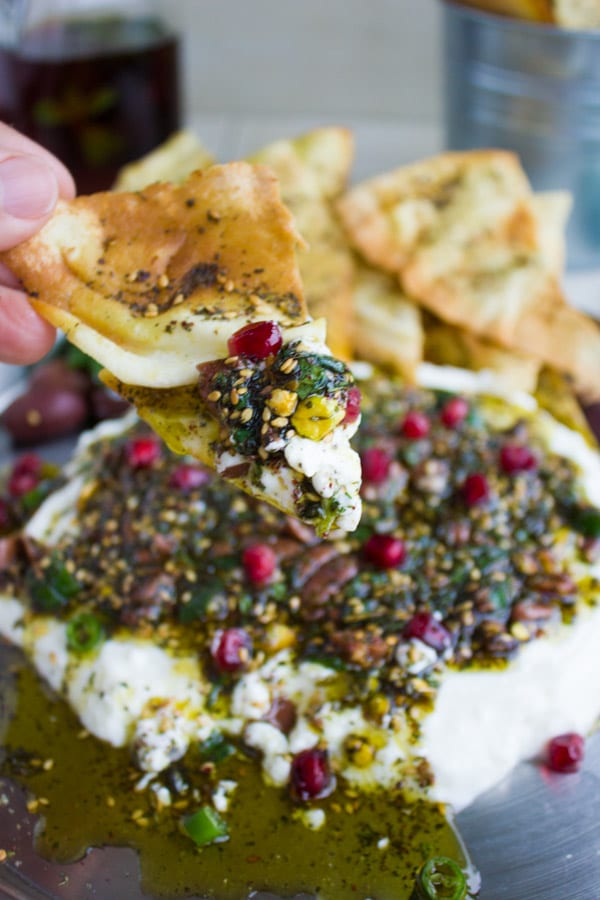 a crispy pita chip just dipped into a creamy white labneh dip topped with zaatar pistachio topping and pomegranate seeds.