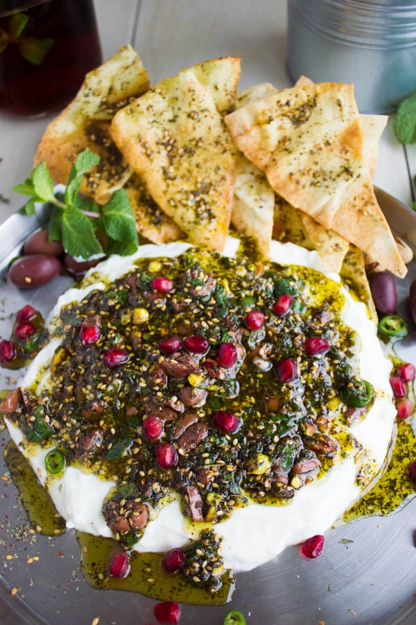 Labneh Dip with Zaatar Pistachio Mint Olive Topping and pomegranate seeds served on a silver platter with a side of zaatar-dusted pita chips and ice tea.