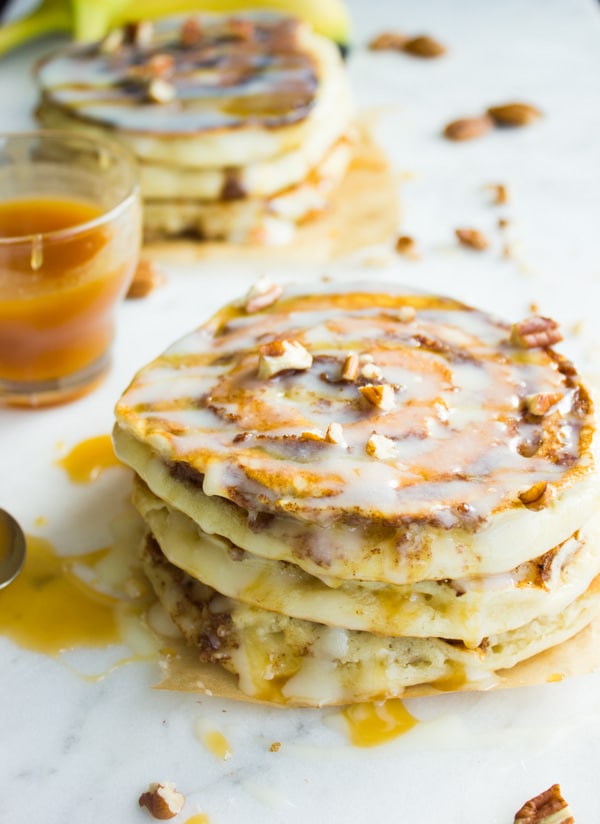 stack of Banana Swirl Cinnamon Roll Pancakes drizzled with cream cheese icing and caramel and sprinkled with chopped pecans