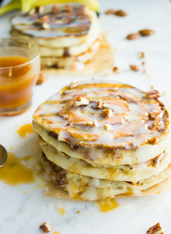  stack of Banana Swirl Cinnamon Roll Pancakes drizzled with cream cheese icing and caramel and sprinkled with chopped pecans