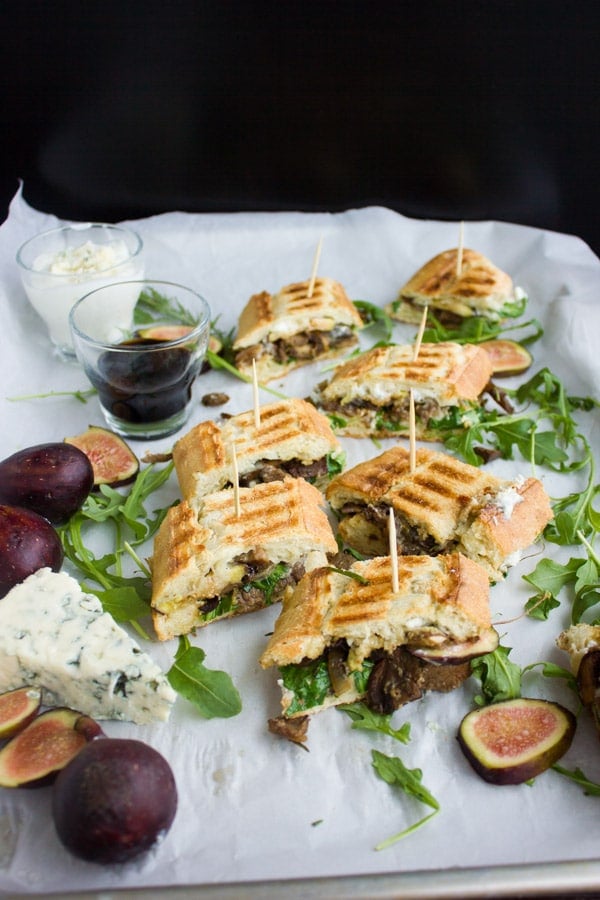 Blue Cheese Philly Steak Sandwich Bites arranged on white parchment paper with some ingredients and a dish with balsamic dip on the side