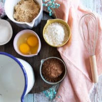 ingredients to make coconut brownies on a board including a bowl of 2 eggs, brown sugar, coconut oil, cocoa powder, salt and flour