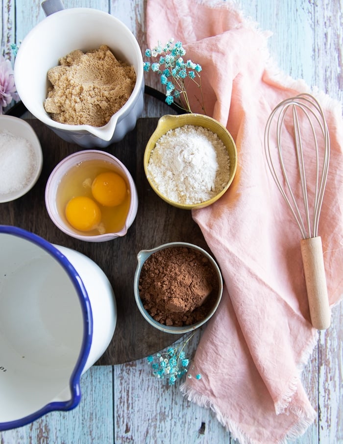 ingredients to make coconut brownies on a board including a bowl of 2 eggs, brown sugar, coconut oil, cocoa powder, salt and flour