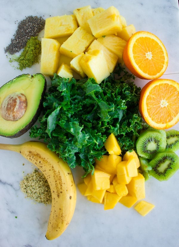 smoothie ingredients like kale, pineapple chunks, kiwi slices, banana, half an avocado and mango cubes piled up on a white surface.