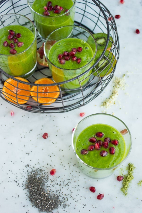 4 glasses with green smoothie decorated with pomegranate seeds on a white tabletop arranged with some tangerines, kiwis and chia seeds