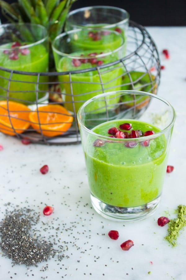 side view of green boosting smoothie sprinkled with pomegranate seeds against the backdrop of a wire basket filled with more smoothie glasses and fruit.