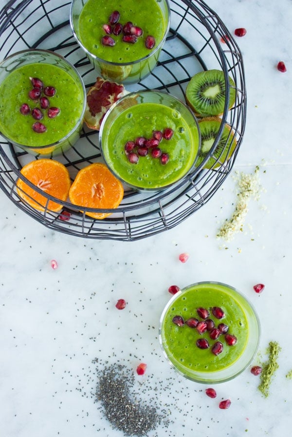 Overhead shot of 4 glasses of green Hemp Matcha Kale Power Boosting Smoothies, three of them in a wire basket with some fruit.