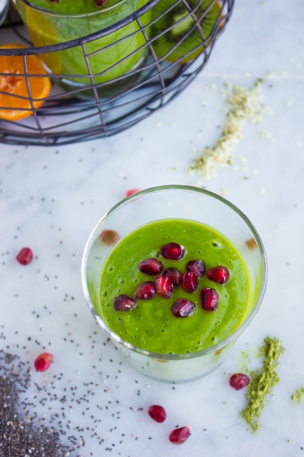 Green Boosting Smoothie on a white tabletop surrounded my some ingredients like hemp seeds, chia seeds and matcha powder.