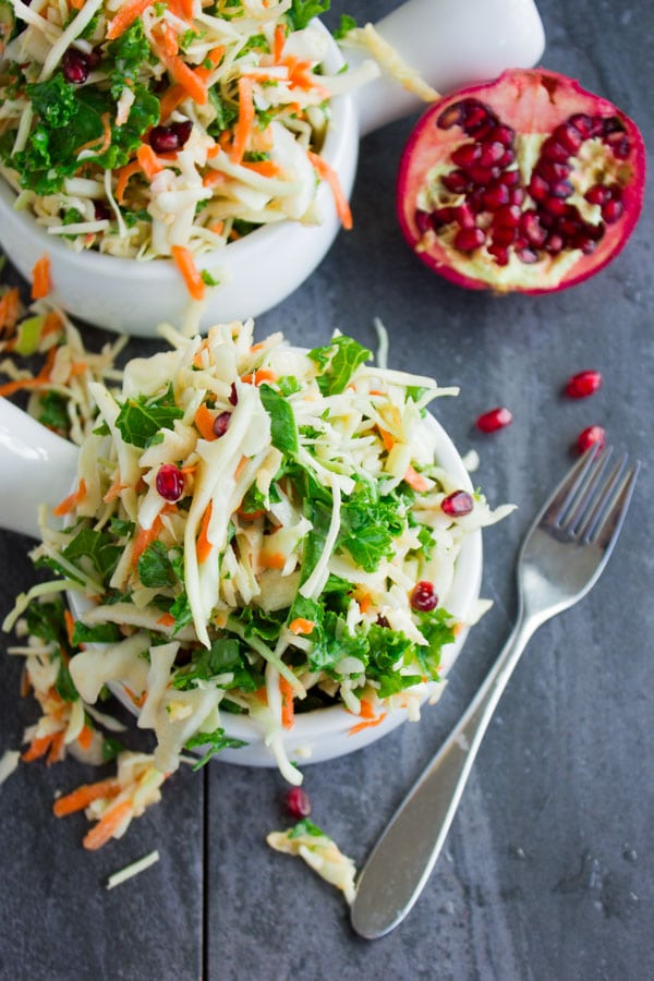 Close-up of a colorful coleslaw with chopped kale, grated apples and pomegranate seeds with half a pomegranate in the background.