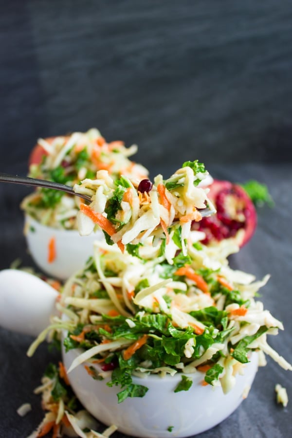 a fork full of coleslaw with kale, grated apples and pomegranate seeds balanced over white bowl filled with more coleslaw.