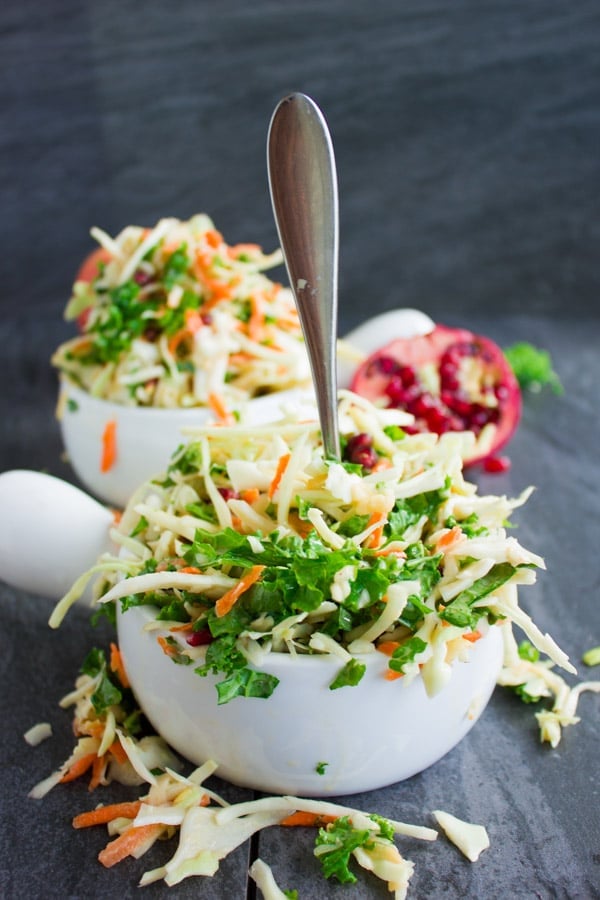 Side view of Coleslaw with Kale, Apples and Pomegranate with a fork sticking out of the front dish and some slaw falling out of the bowl onto the table. 