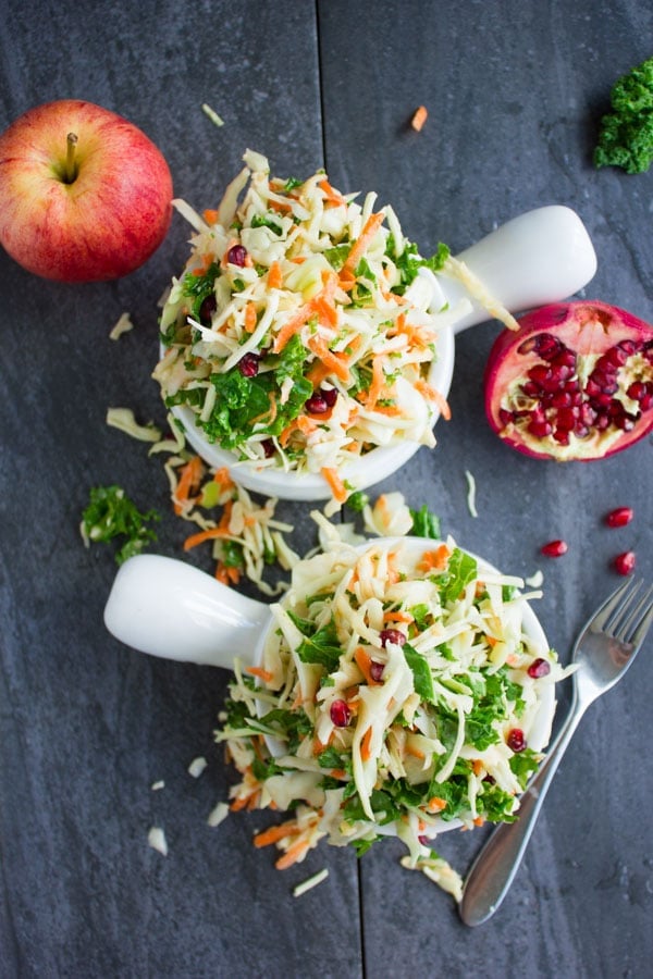 Overhead shot of two bowls of Coleslaw with kale, grated apples and carrots and sprinkled with pomegranate seeds on a rustic dark table.