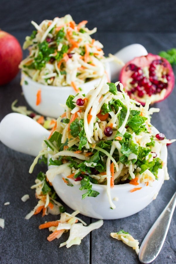 Coleslaw with kale, shredded apples and pomegranate seeds piled up in two white serving bowls on a black rustic table.