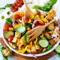 A hand adding all panzanella salad ingredients into a salad bowl and using a spoon to toss them