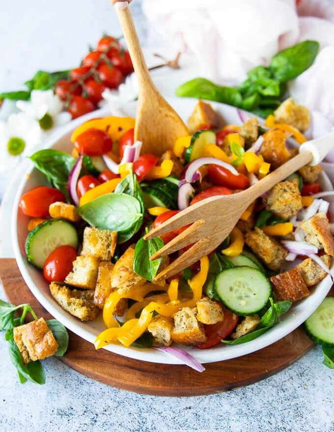 A hand adding all panzanella salad ingredients into a salad bowl and using a spoon to toss them 