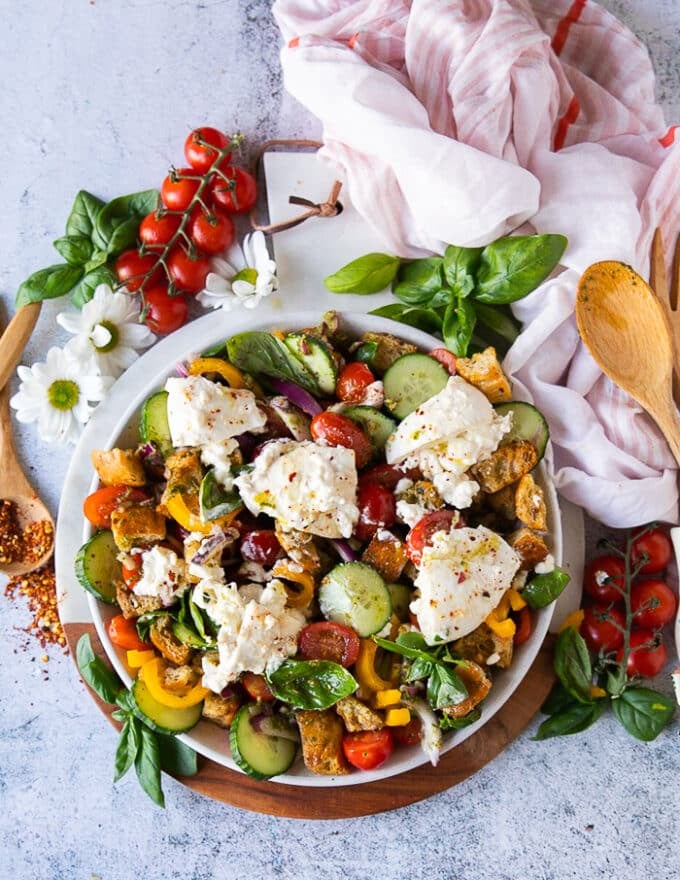 Panzanella salad over a white board surrounded by baby tomatoes, chunks of croutons, lots of fresh basil leaves, some chilli flakes in a bowl and a tea towel