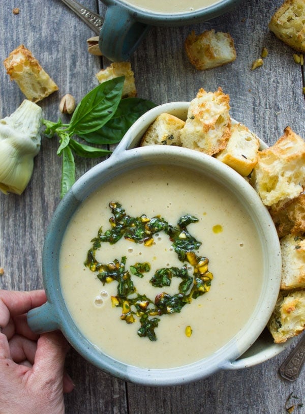 overhead shot of a bowl of creamy artichoke soup topped with pistachio pesto surrounded by croutons