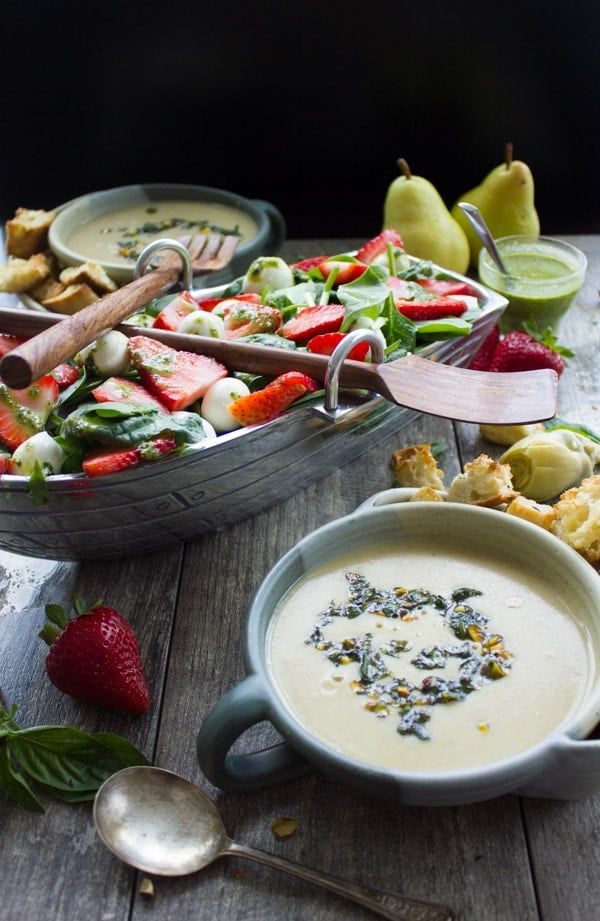 a bowl with pesto-topped artichoke soup in front of a boat-shaped bowl filled with Strawberry Spinach Salad.