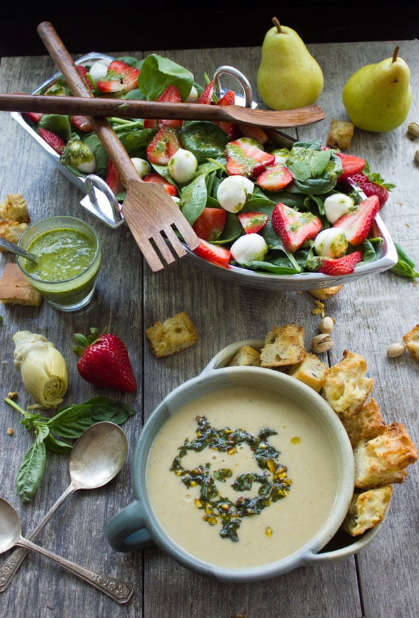 a bowl with pesto-topped artichoke soup in front of a boat-shaped bowl filled with Strawberry Spinach Salad.