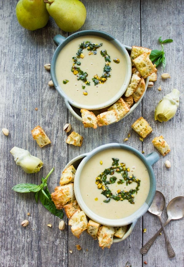 overhead shot of two blue bowls of creamy artichoke soup topped with pistachio pesto surrounded by croutons