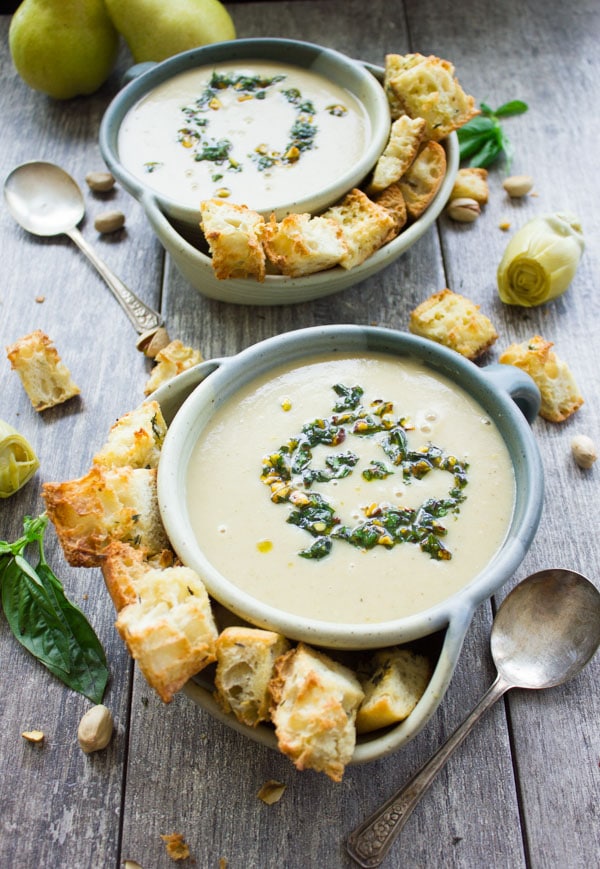 overhead shot of two blue bowls of creamy artichoke soup topped with pistachio pesto surrounded by croutons