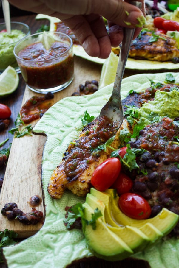 salsa being drizzled on top of spice crusted pan-fried tilapia fillets with black beans and guacamole on the side