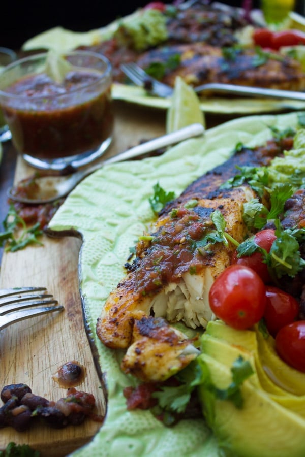 close up of Mexican pan-fried tilapia fillet crusted with spices served next to sliced avocado and a dish with homemade salsa