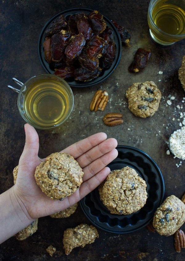 a hand holding a Vegan Oatmeal Raisin Cookie with ingredients and tea mugs in the background