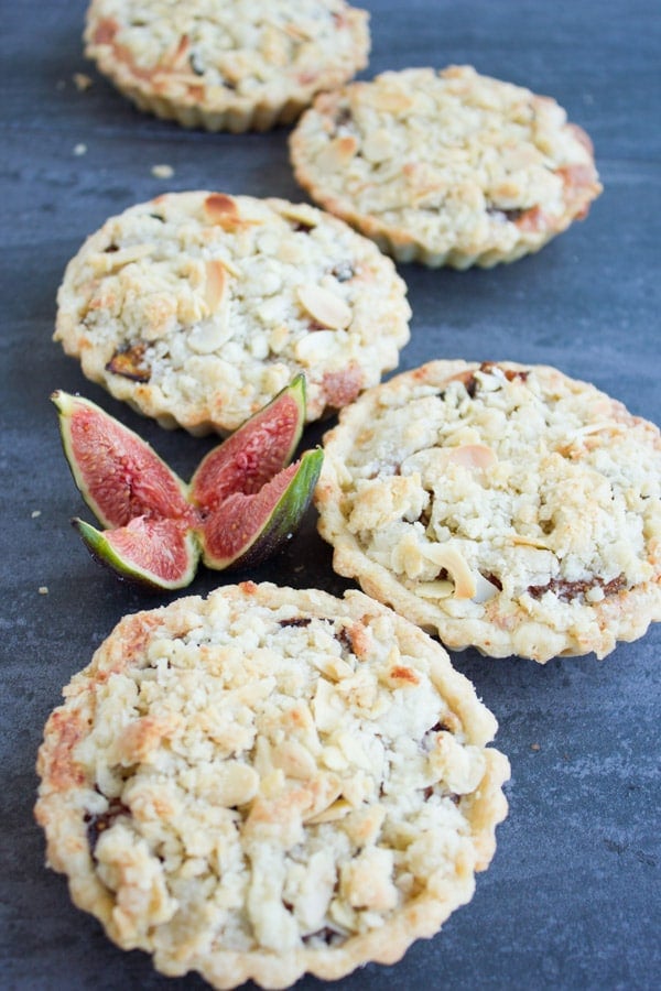 Individual Fig Almond Frangipane Tarts arranged on a rustic dark tabletop with a quartered fresh fig in the background