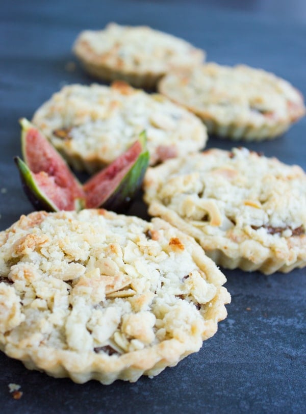 Individual Fig Almond Frangipane Tarts arranged on a rustic dark tabletop with a quartered fresh fig in the background