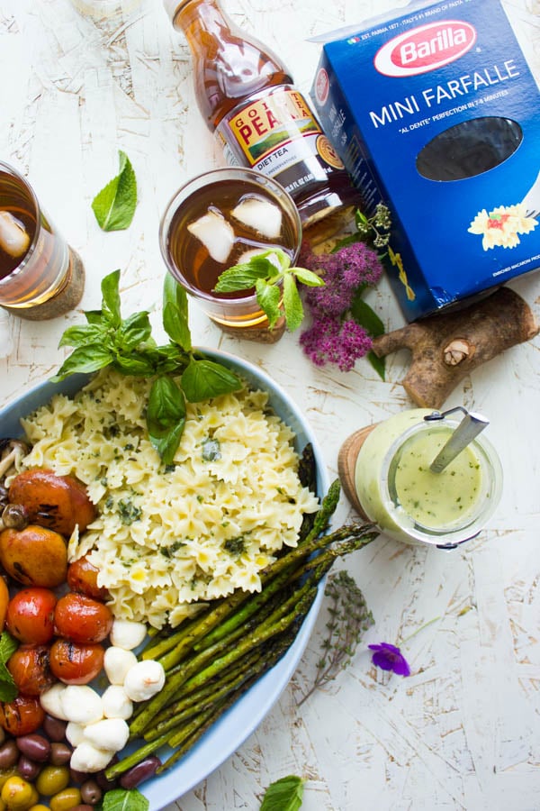 Overhead shot of summer pasta salad plated in an oval dish with grilled vegetables, asparagus and mini mozzarella. Dressing on the side and glasses of sweet tea in the background.