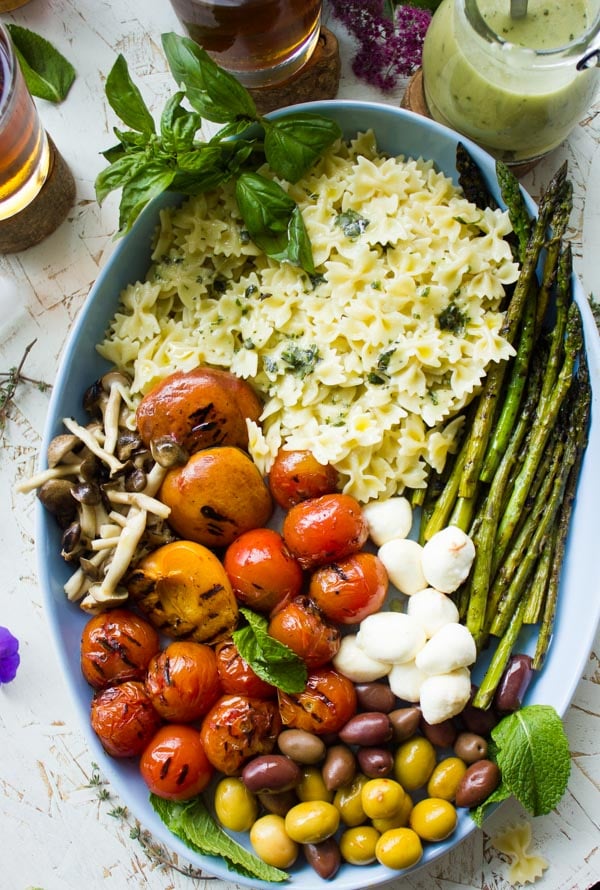 Overhead shot of summer pasta salad plated in an oval dish with grilled vegetables, asparagus, olives and mini mozzarella. Dressing on the side and glasses of sweet tea in the background.