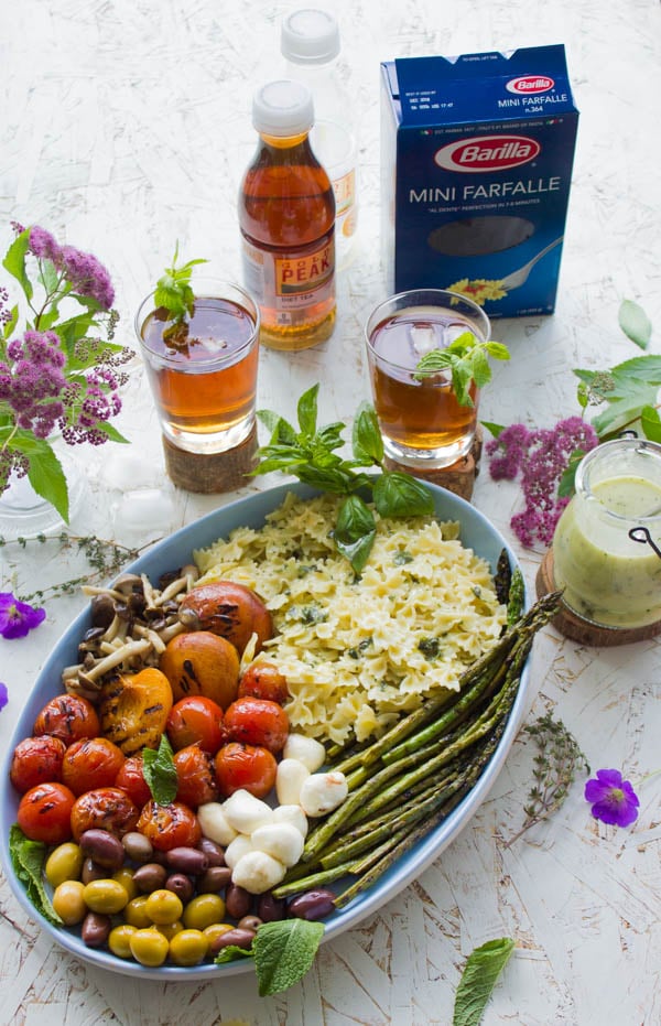 Overhead shot of summer pasta salad plated in an oval dish with grilled vegetables, asparagus and mini mozzarella. Dressing on the side and glasses of sweet tea in the background.