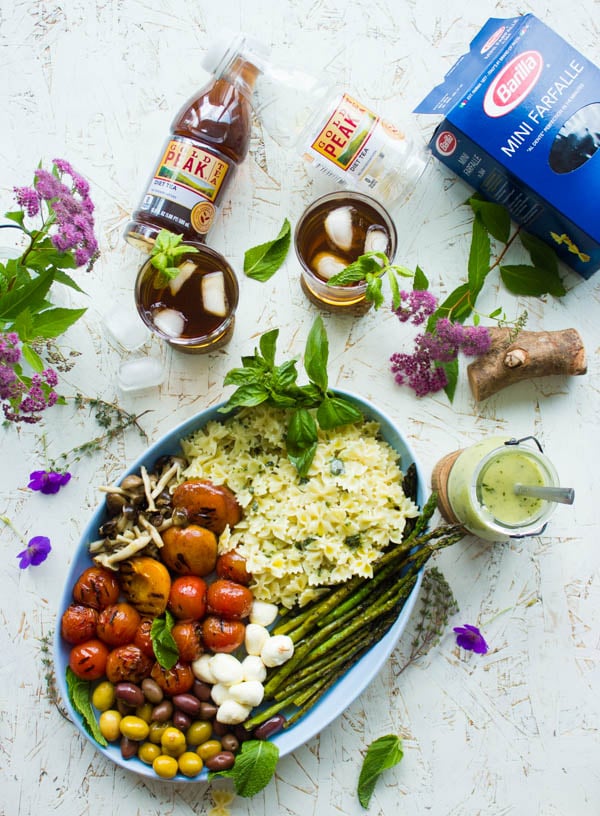 Overhead shot of summer pasta salad plated in an oval dish with grilled vegetables, asparagus and mini mozzarella. Dressing on the side and glasses of sweet tea in the background.